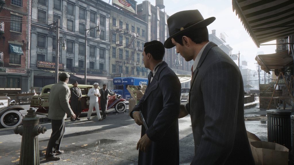 Two men in suits and hats speaking on a city street in Mafia: Definitive Edition.